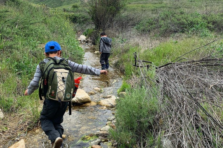 Two boys walking in a creek along a trail with hills in the background.