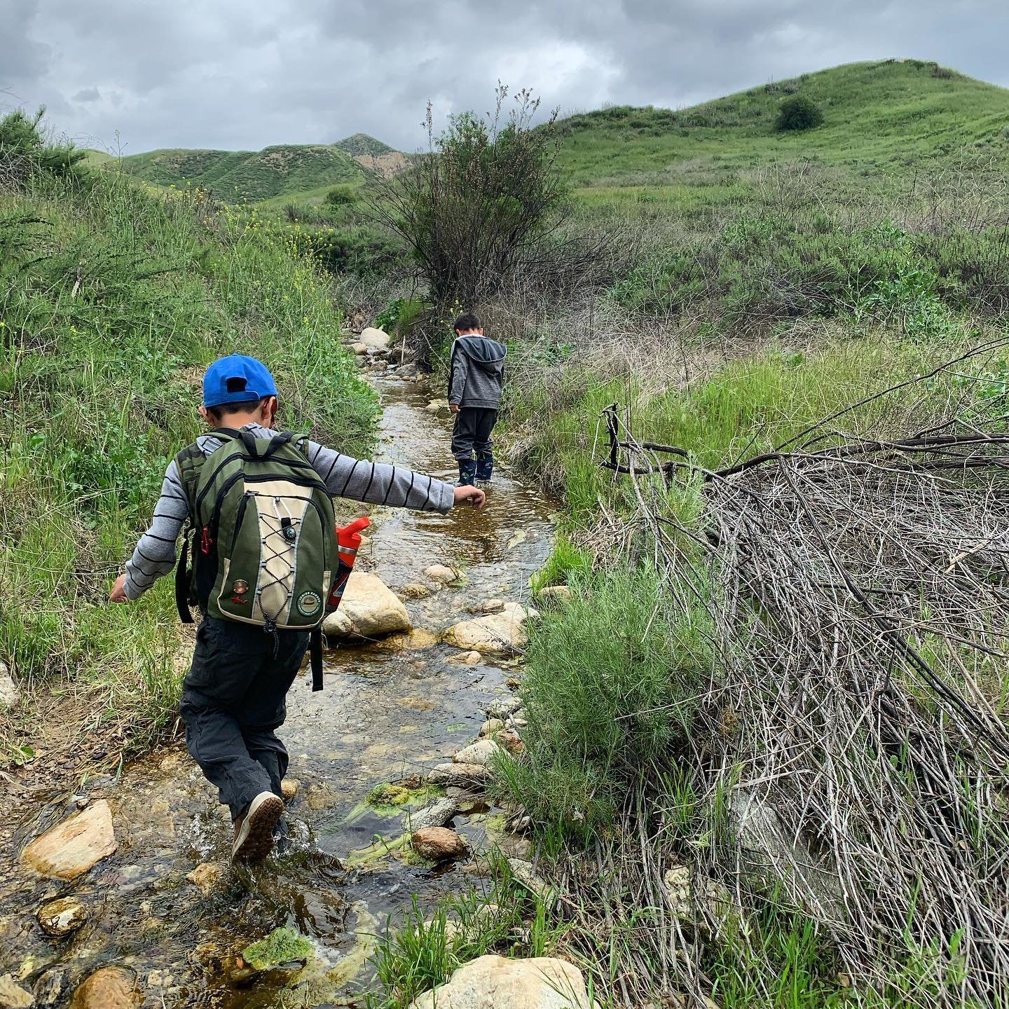 Two boys walking in a creek along a trail with hills in the background.