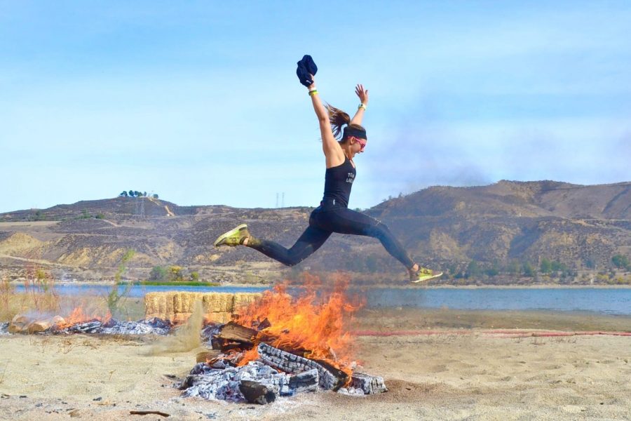 Women jumping over a lit campfire at the Los Angelos Spartan Event Weekend outside near a lake.