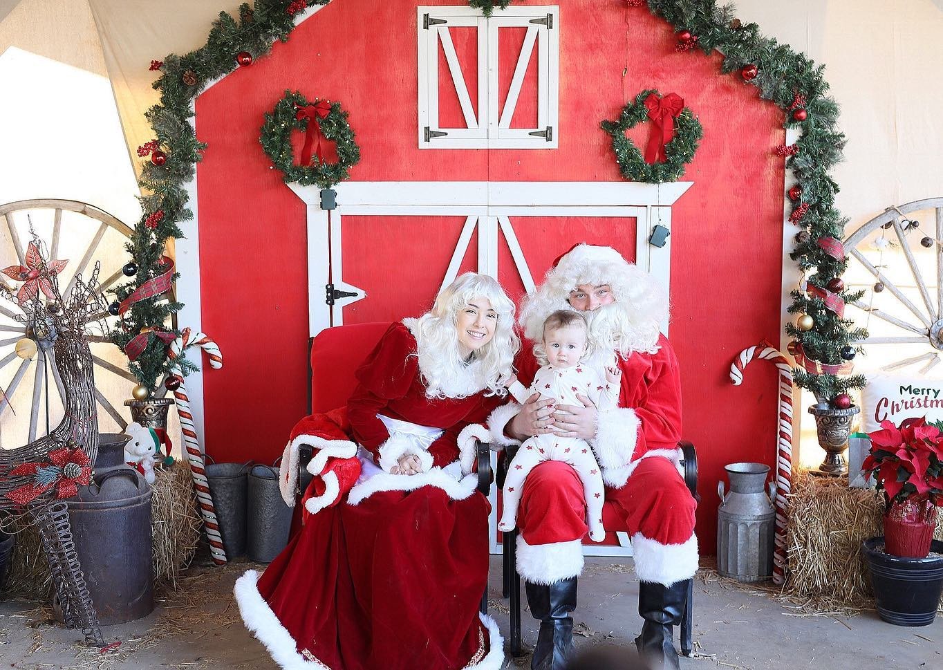 Santa and Mrs. Claus holding a baby in front of a red barn at Gilchrist Farm in Santa Clarita.